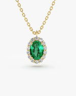 A close-up of a gold chain necklace with an oval-cut emerald green gemstone pendant, which is surrounded by a halo of small, round-cut clear gemstones.