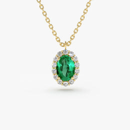 A close-up of a gold chain necklace with an oval-cut emerald green gemstone pendant, which is surrounded by a halo of small, round-cut clear gemstones.