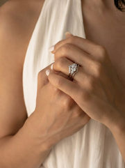 A close-up of a two-piece silver ring set held between fingers, showing a main ring with a large central clear stone and a contoured band with smaller accent stones.

