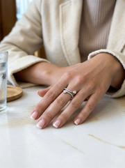A close-up view of a hand holding a silver open-style ring. The ring features a cluster of small, clear, sparkling gemstones in the center, with more small stones lining the two overlapping bands.